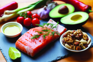 Foods high in healthy fats such as Salmon, olive oil, nuts and avocados with vegetables and herbs on table top.